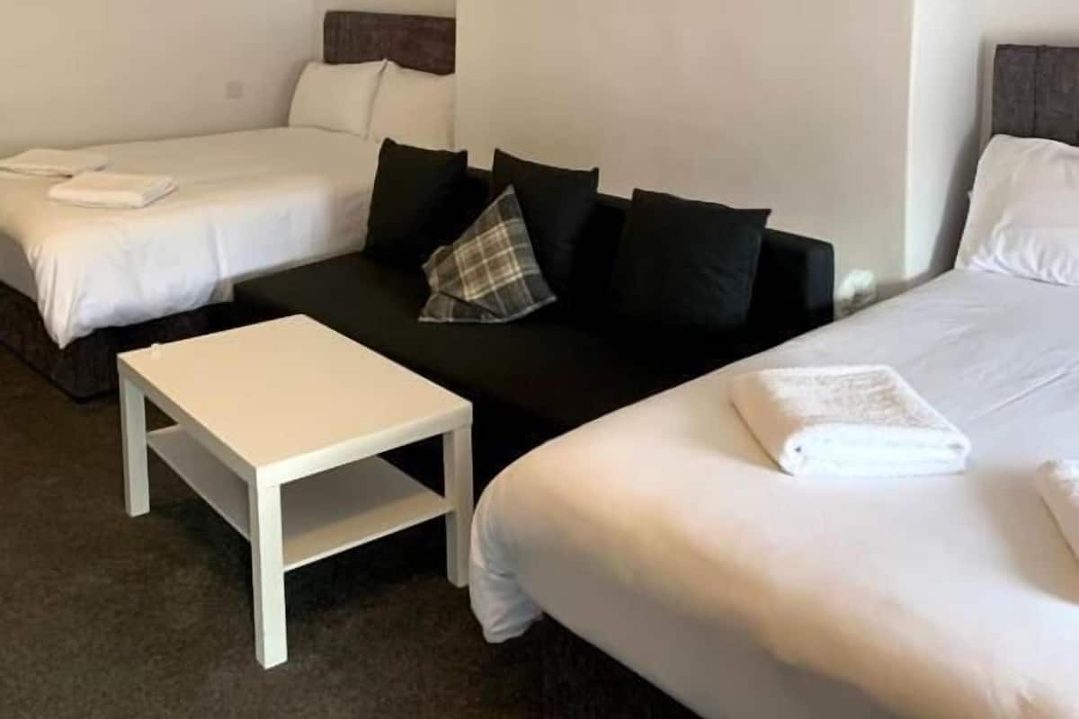 Habitación de hotel con dos camas individuales, un sofá cama, una mesita pequeña y una lámpara en el techo.