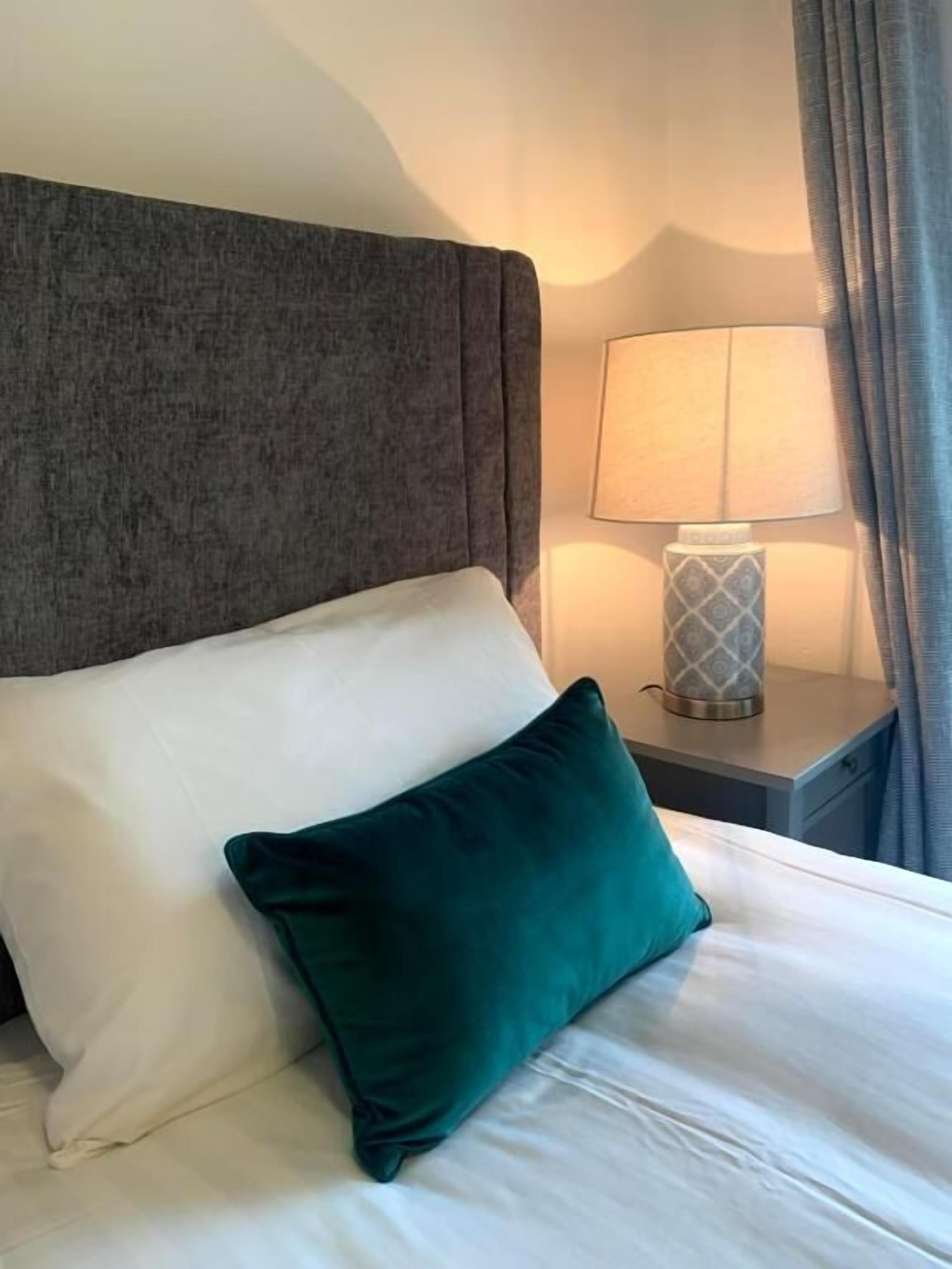 Una cama bien hecha con un cojín blanco y verde azulado, un cabecero gris y una lámpara de noche en una mesita.