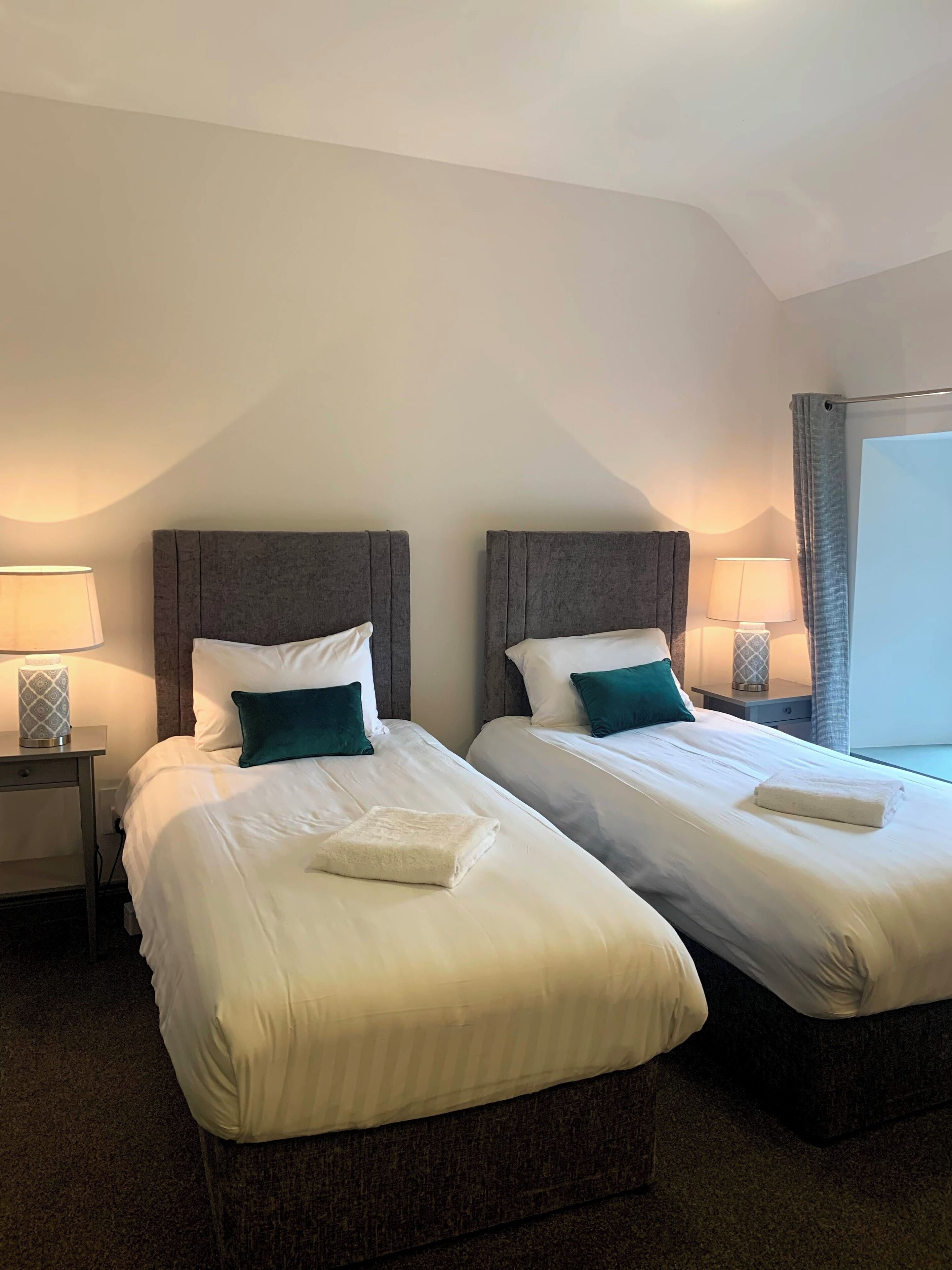 Habitación de hotel con dos camas, cada una con ropa blanca y almohadas turquesa, una mesita de noche con lámpara y una ventana con cortinas.