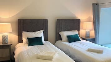Habitación de hotel con dos camas, cada una con ropa blanca y almohadas turquesa, una mesita de noche con lámpara y una ventana con cortinas.