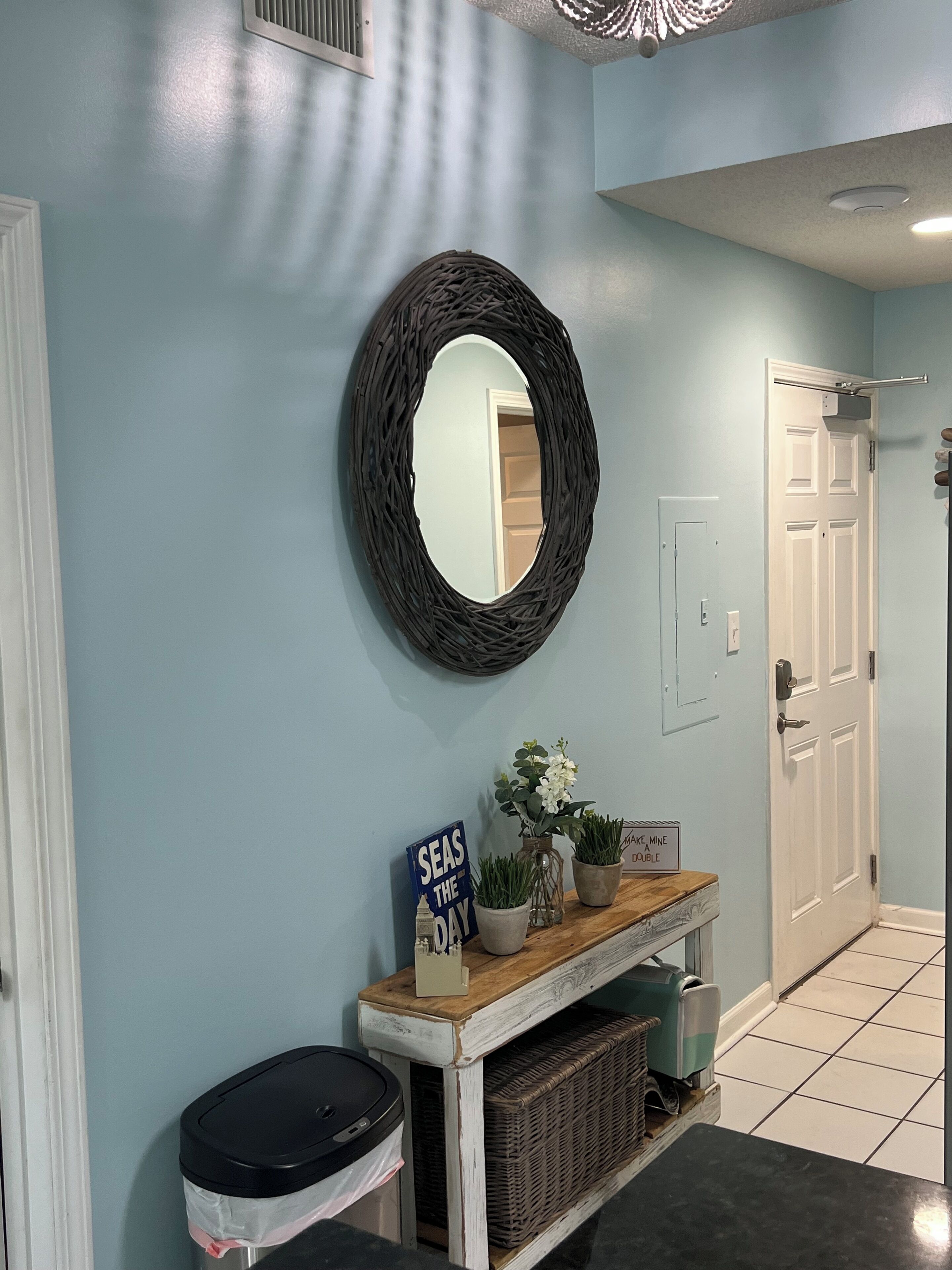 A hallway with a light blue wall, a round mirror, a wooden console table with a sign, a trash bin, and a door.