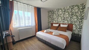 A hotel room with a bed, a radiator, a window with curtains, and a wall with a leafy pattern. - Quiet apartment Vittel - Near the thermal baths (Norroy)