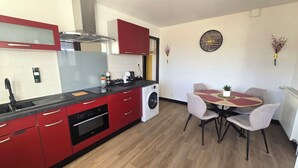 A compact kitchen with red cabinets, a built-in oven, and a washing machine. There is a dining table with chairs and a clock on the wall. - Quiet apartment Vittel - Near the thermal baths (Norroy)