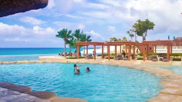 Zona de piscina con agua azul cristalina, una formación rocosa y un pabellón con cerca blanca.