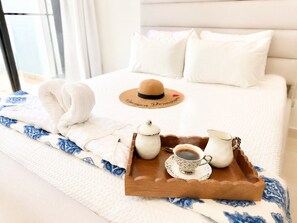 Un lit bien fait, recouvert d’une couette blanche et d’un plaid à motifs bleus et blancs. Un plateau en bois, sur lequel se trouvent une théière et d’autres éléments de service à thé, ainsi qu’un chapeau, est posé sur le lit.