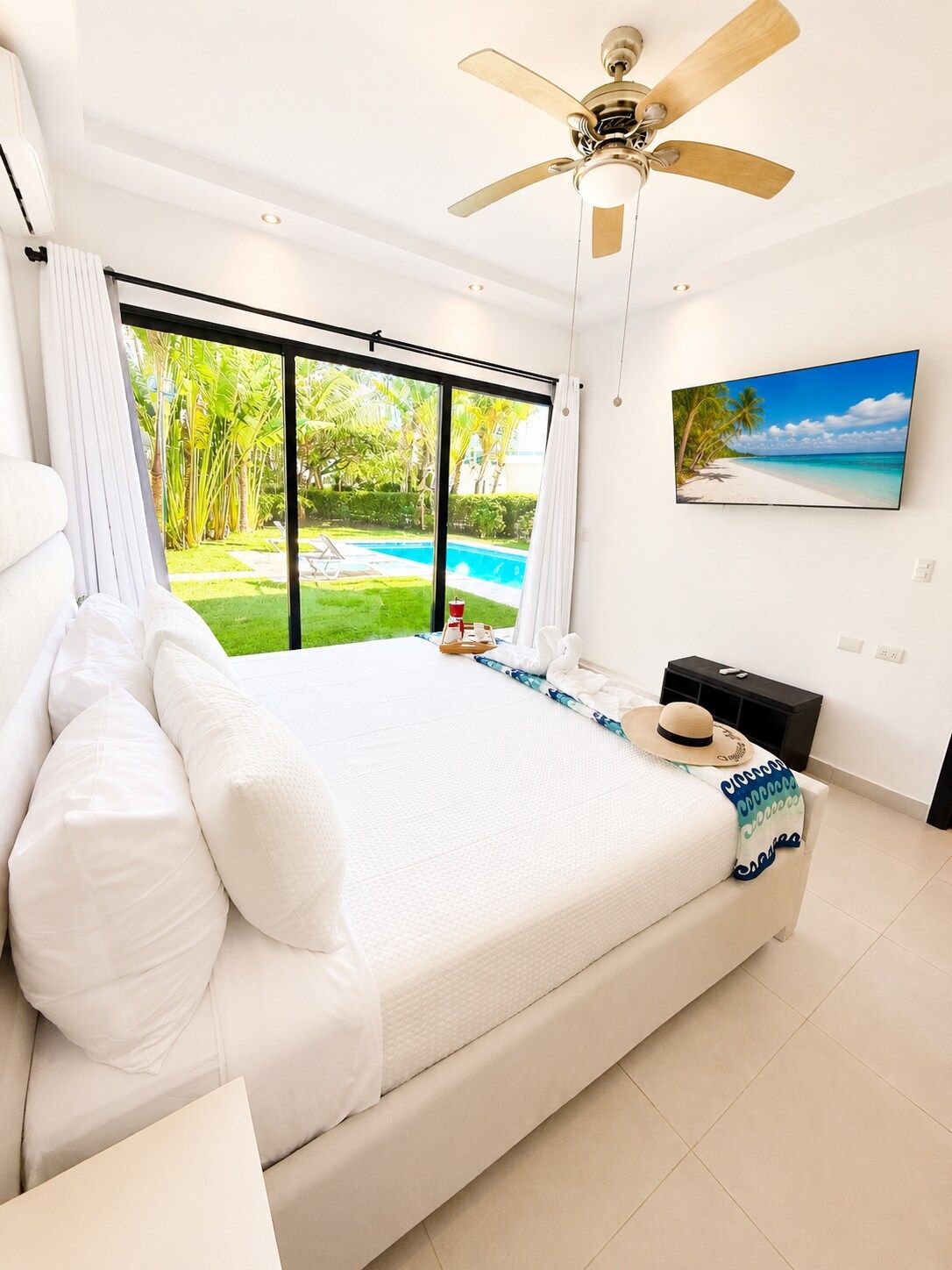 Een slaapkamer met een groot bed, een plafondventilator, uitzicht op een zwembad en een stranddecoratie aan de muur.