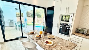 Una cocina moderna con una isla central, taburetes de bar, horno integrado y vistas a un jardín con piscina.