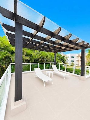 A covered outdoor area with two white lounge chairs and a small table, surrounded by a glass railing and a view of greenery and buildings. - Villa Serena| 4 Suites, Pool, Terrace & Corner Lot (Sosúa)