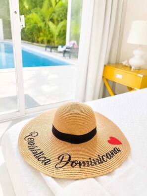 A straw hat with embroidered text on a white surface, a yellow bedside table with a lamp, and a view of a swimming pool through a window. - Villa Serena| 4 Suites, Pool, Terrace & Corner Lot (Sosúa)