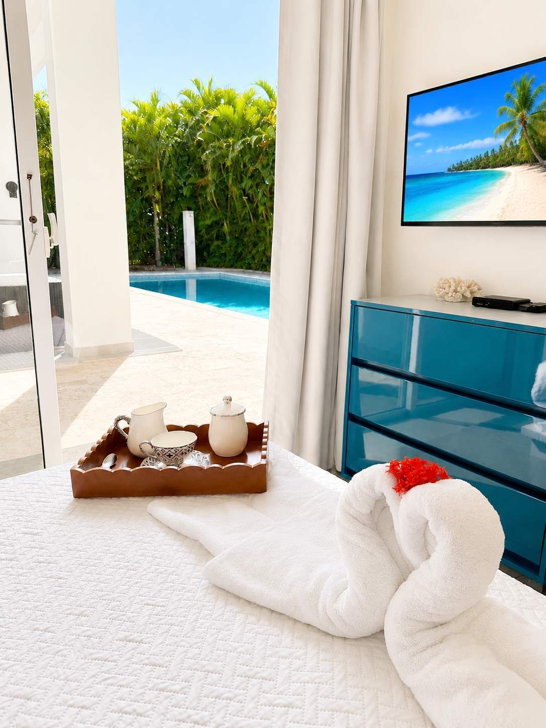 Ein Hotelzimmer mit Bett, Tablett mit Teeservice, Handtuch, Fernseher an der Wand und Blick auf Pool und Palmen.
