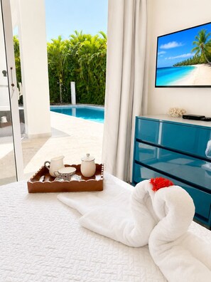 A hotel room with a bed, a tray with tea set, a TV displaying a beach scene, and a view of a pool and greenery. - Villa Serena| 4 Suites, Pool, Terrace & Corner Lot (Sosúa)