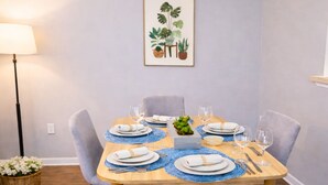 Un coin repas avec une table en bois dressée pour quatre personnes, entourée de chaises grises, d’une petite plante en pot et d’une illustration botanique encadrée accrochée au mur.