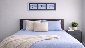 Un lit avec une couette bleue, des oreillers et un plaid beige. Il y a un tableau encadré au-dessus du lit et une petite table avec une plante et une tasse sur le côté.