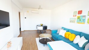 A modern living room with a blue sofa, a glass coffee table, a white TV stand, and a dining area with a small table and chairs. - One-bedroom seafront apartment on Mackenzie with stunning sea views. (Larnaca)