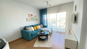 A living room with a teal sofa, a glass coffee table, and a large window with blue curtains. - One-bedroom seafront apartment on Mackenzie with stunning sea views. (Larnaca)