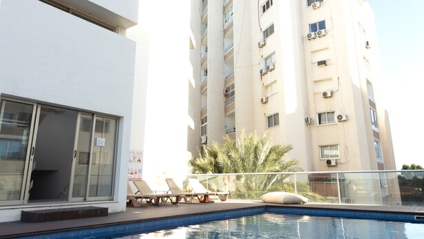 A swimming pool with a wooden deck, a white building with balconies, and a clear sky. - One-bedroom seafront apartment on Mackenzie with stunning sea views. (Larnaca)