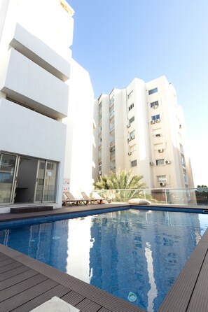 Una piscina con un pontile in legno, un edificio bianco con balconi e un cielo sereno.