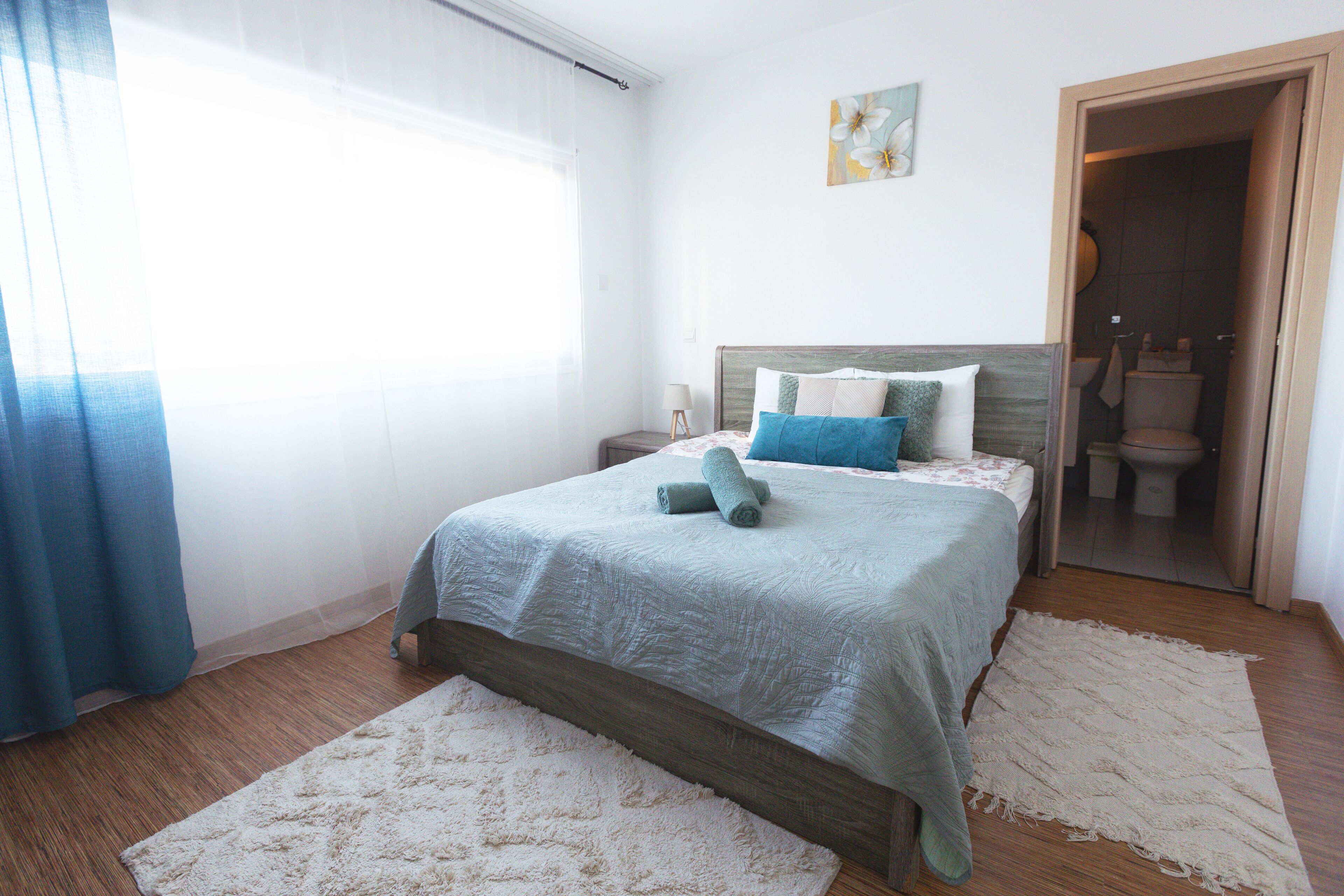A bedroom with a bed, a nightstand, a window with curtains, a painting on the wall, and an open door to the bathroom.