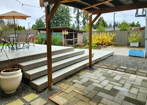 A covered outdoor patio with steps leading to a seating area, surrounded by a wooden fence and various plants. - A Rather Posh 4 BR Renton Retreat with AC (Renton)