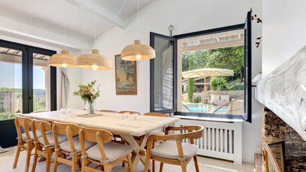 Ein Essbereich mit einem großen Holztisch, sechs Stühlen und hängenden Korbtabletten. Ein großes Fenster bietet Blick auf einen Außenpool und Garten.