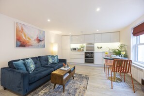 A modern living room with a dark blue sofa, a wooden coffee table, and a kitchen in the background. - Stylish 1-Bed Apartment in Fairview (Cheltenham)