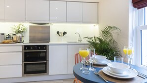 A modern kitchen with white cabinets, a built-in oven, a sink, and a dining area with a table set for two. - Stylish 1-Bed Apartment in Fairview (Cheltenham)