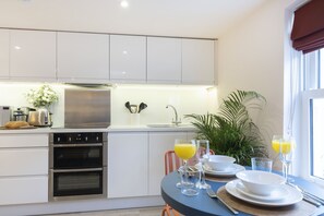 A modern kitchen with white cabinets, a built-in oven, a sink, and a dining area with a table set for two. - Stylish 1-Bed Apartment in Fairview (Cheltenham)