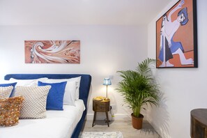 A bedroom with a bed, pillows, a nightstand with a lamp, a potted plant, and two artworks on the wall. - Stylish 1-Bed Apartment in Fairview (Cheltenham)