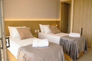 Ein Hotelzimmer mit zwei Betten, jeweils mit weißen Laken und einem braunen Zierkissen. Es gibt ein Holz-Kopfteil, einen Nachttisch mit einer Lampe und eine Tür zu einem weiteren Raum.
