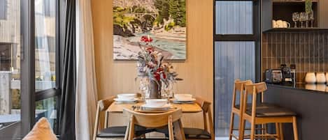 A dining area with a table set for two, a painting of a landscape, and a bar area with coffee machines.