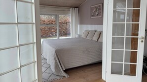 A bedroom with a large bed, a door with glass panels, a window with curtains, and a ceiling with wooden beams. - Spacious holiday home near beach and nature (Jægerspris)