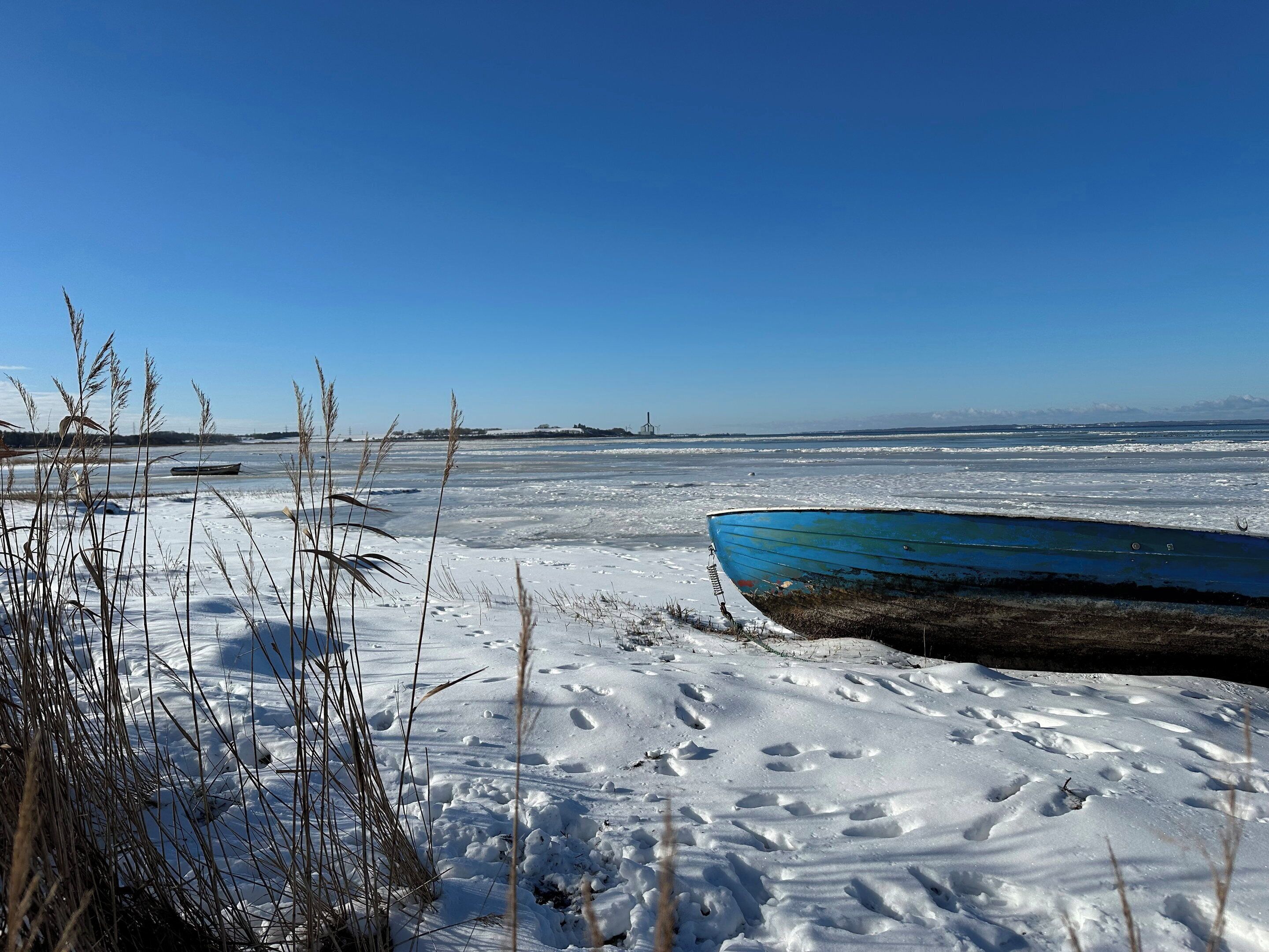 Un paysage hivernal, avec un bateau bleu sur la glace, un sol recouvert de neige et une végétation clairsemée.
