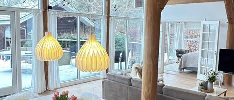 A spacious room with a large dining table, wooden beams, and large windows offering a view of the outdoors.