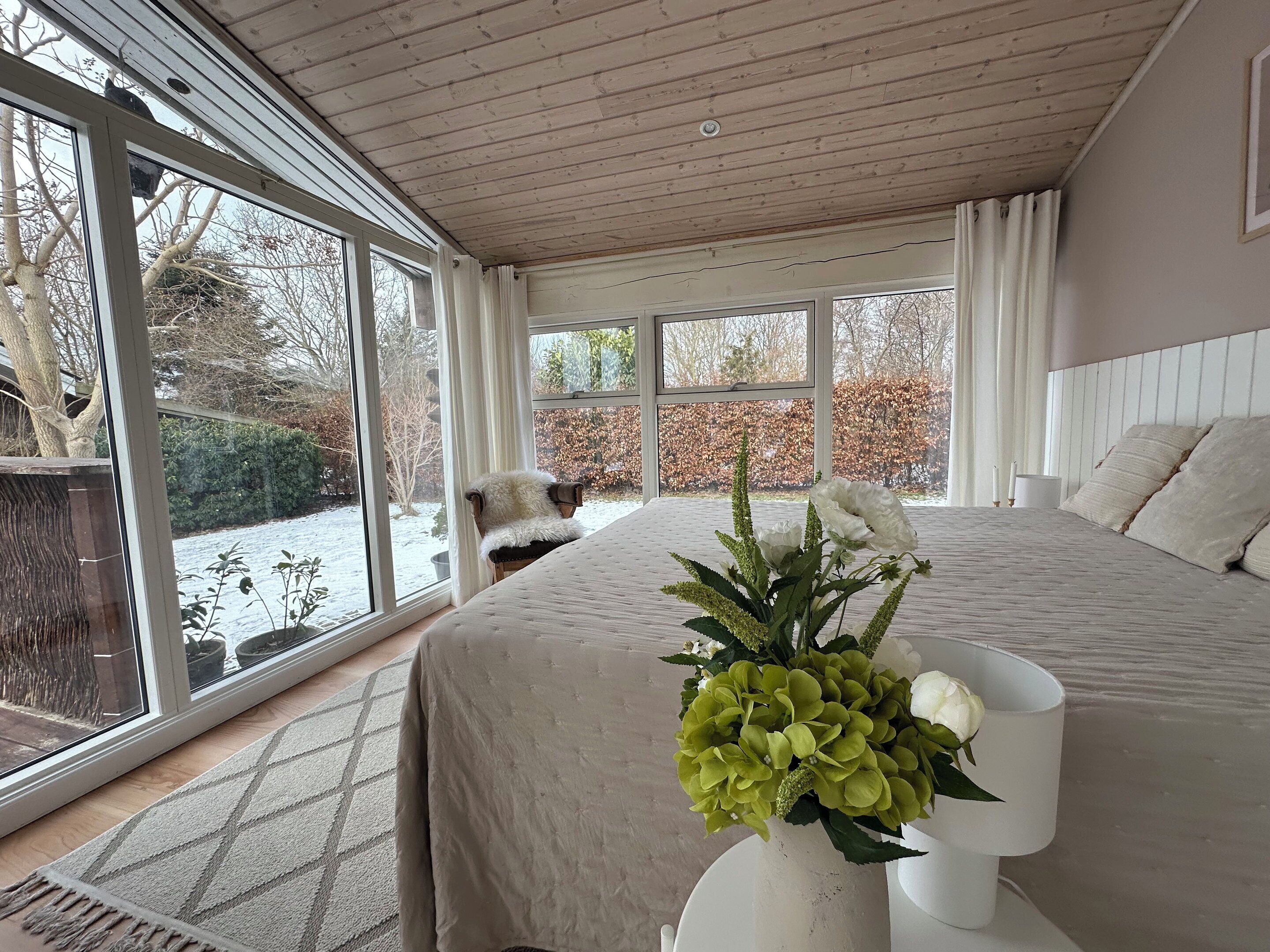A bedroom with a large window, a bed with a white duvet, a vase with flowers, and a chair by the window.