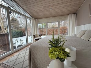 Een slaapkamer met een groot raam, een bed met een witte dekbedovertrek, een vaas met bloemen en een stoel bij het raam.
