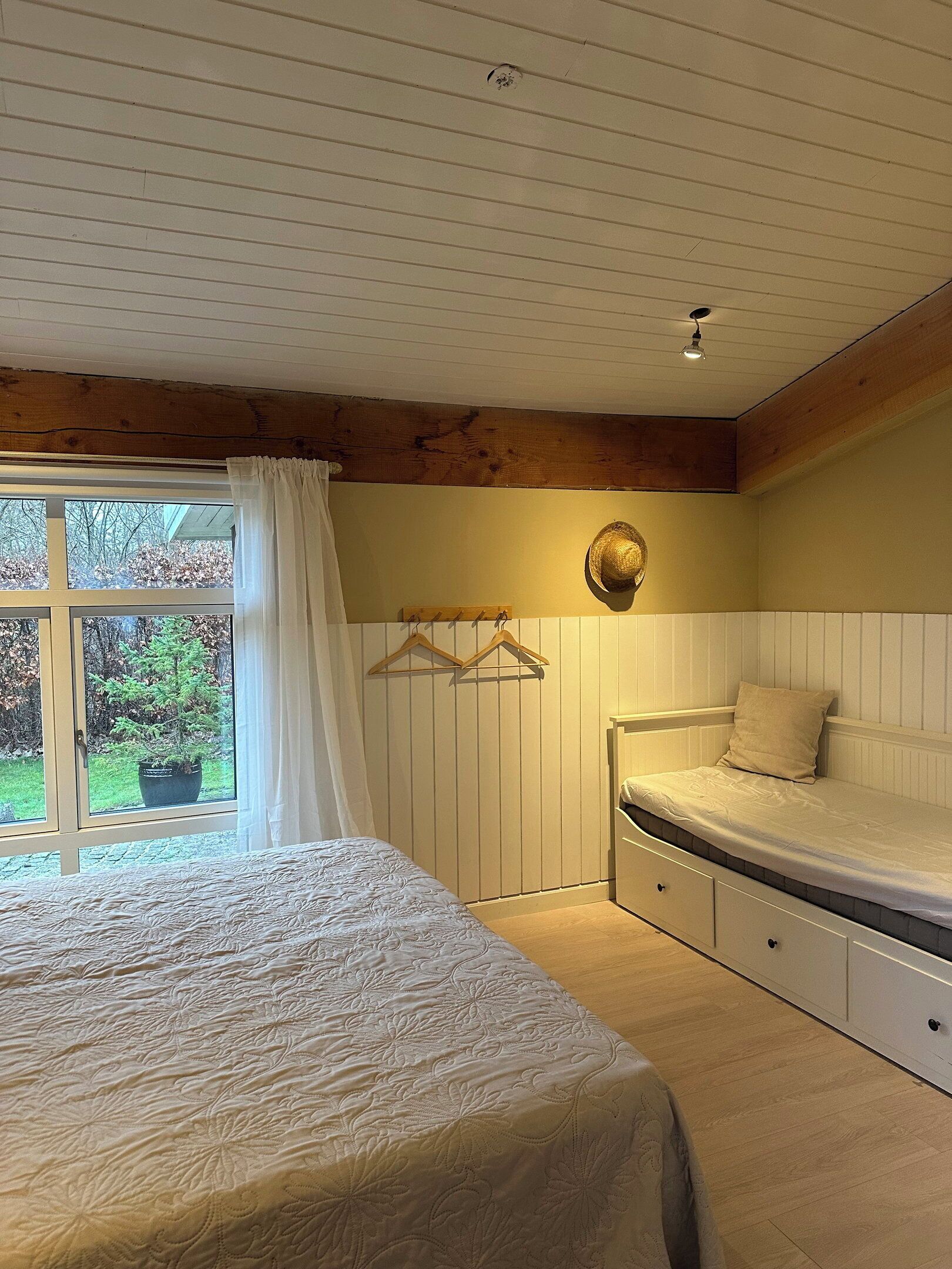 Un dormitorio con cama, mesita de noche, un sombrero colgado, ventana con cortinas y vistas al exterior.
