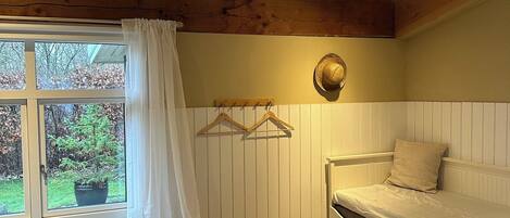 A bedroom with a bed, a bedside table, a hanging hat, a window with curtains, and a view of the outdoors.