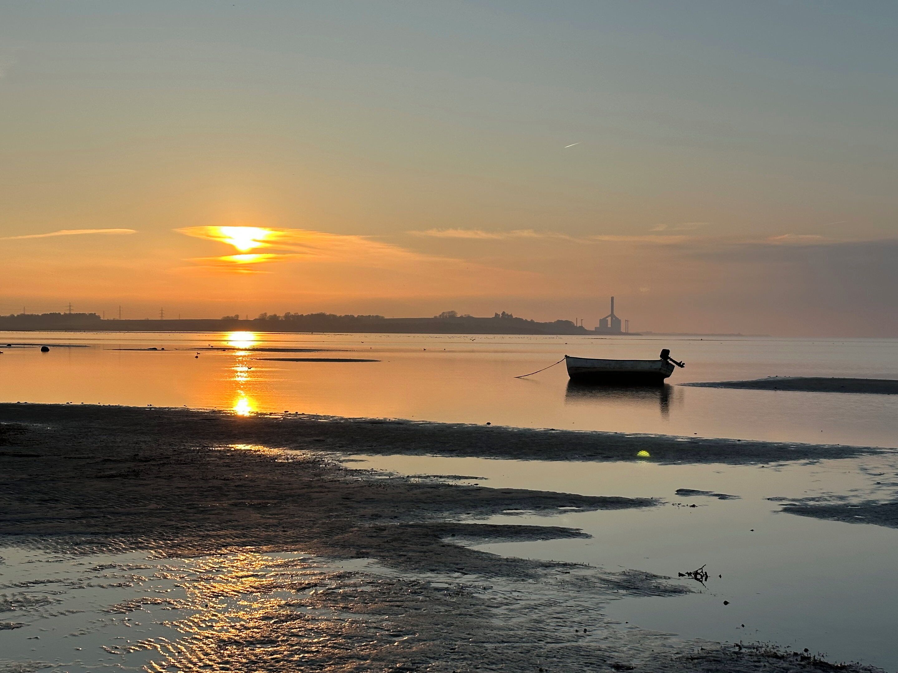 A calm body of water with a boat anchored near the shore, a distant industrial structure, and a clear sky.