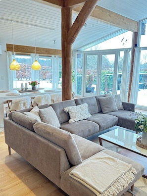 A modern living room with a sectional sofa, a glass coffee table, and large windows offering a view of the outdoors.