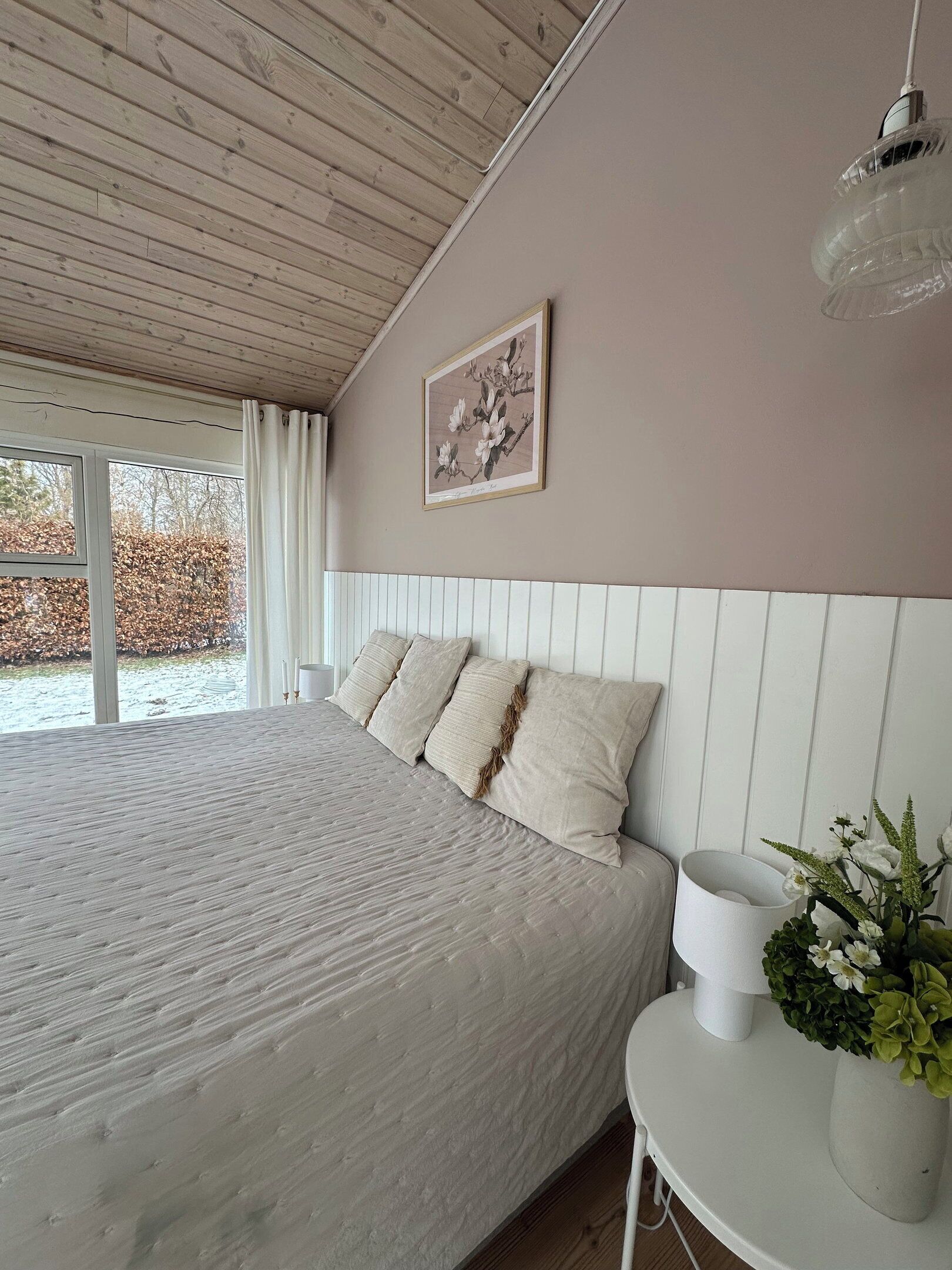 A bedroom with a large bed, a bedside table with a lamp, a framed picture on the wall, and a view of the outdoors.