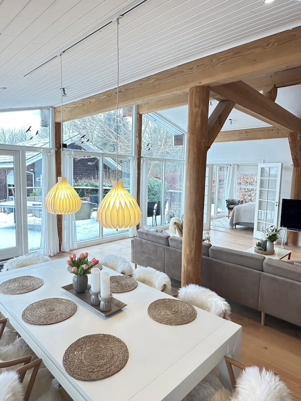 A spacious room with a large dining table, wooden beams, and large windows offering a view of the outdoors.