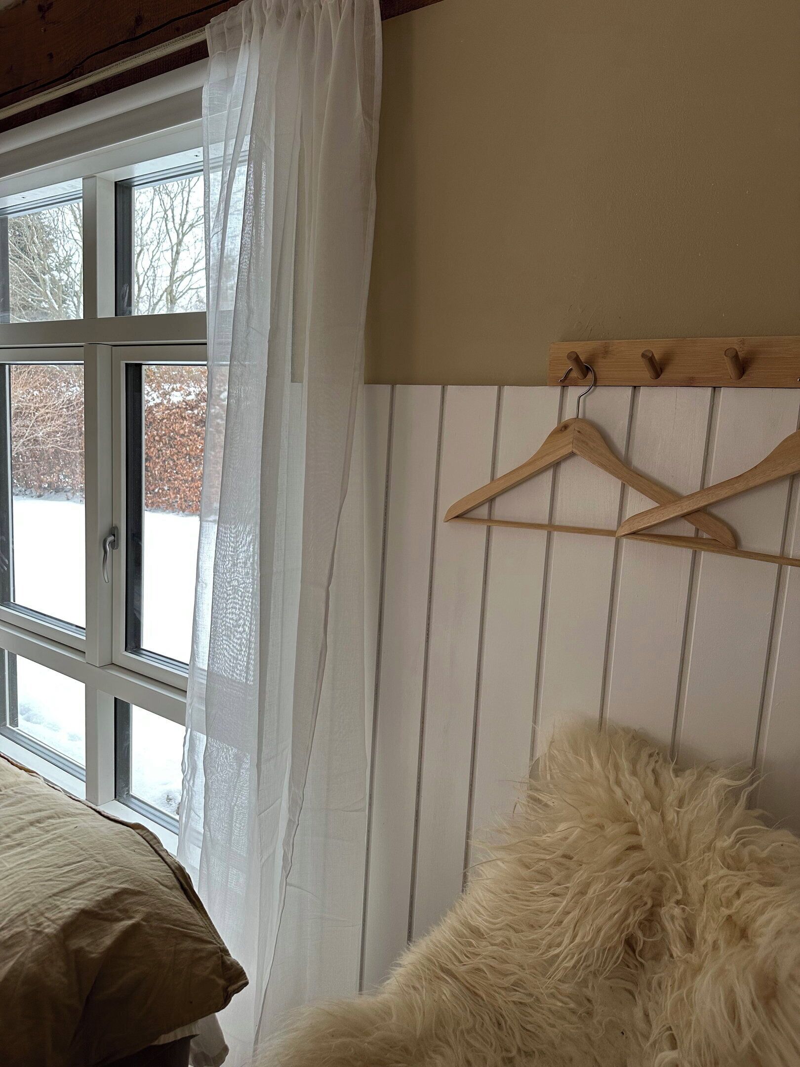 Une chambre avec un lit, un porte-manteau en bois muni de cintres, une fenêtre donnant sur des arbres et un rideau blanc.
