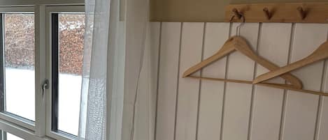 Une chambre avec un lit, un porte-manteau en bois muni de cintres, une fenêtre donnant sur des arbres et un rideau blanc.