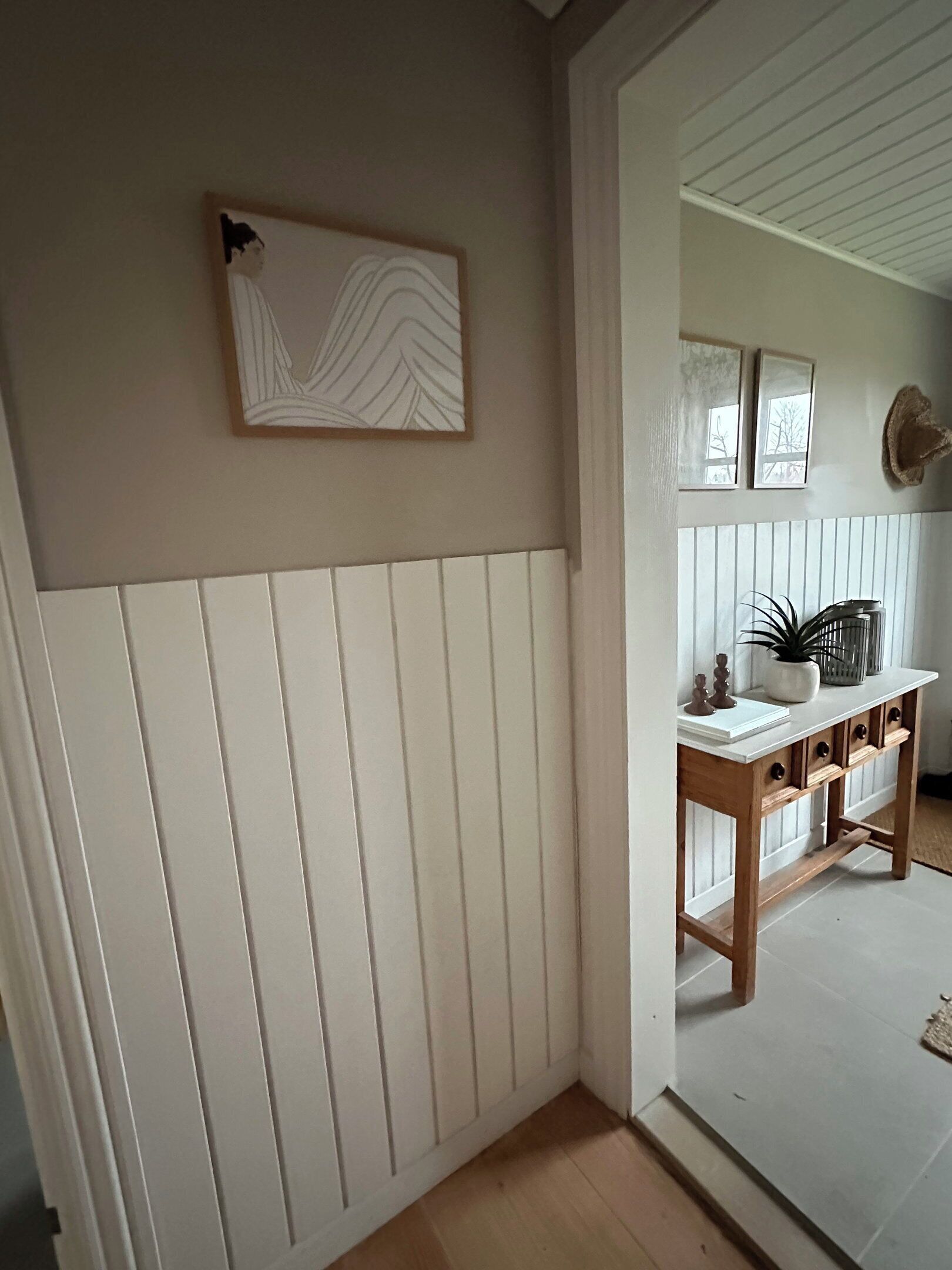 Un couloir avec un mur recouvert de panneaux blancs, une console en bois ornée d’objets décoratifs et une œuvre d’art encadrée.