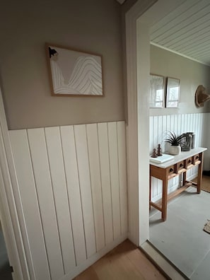 Un pasillo con pared blanca panelada, una consola de madera con objetos decorativos y una obra de arte enmarcada.