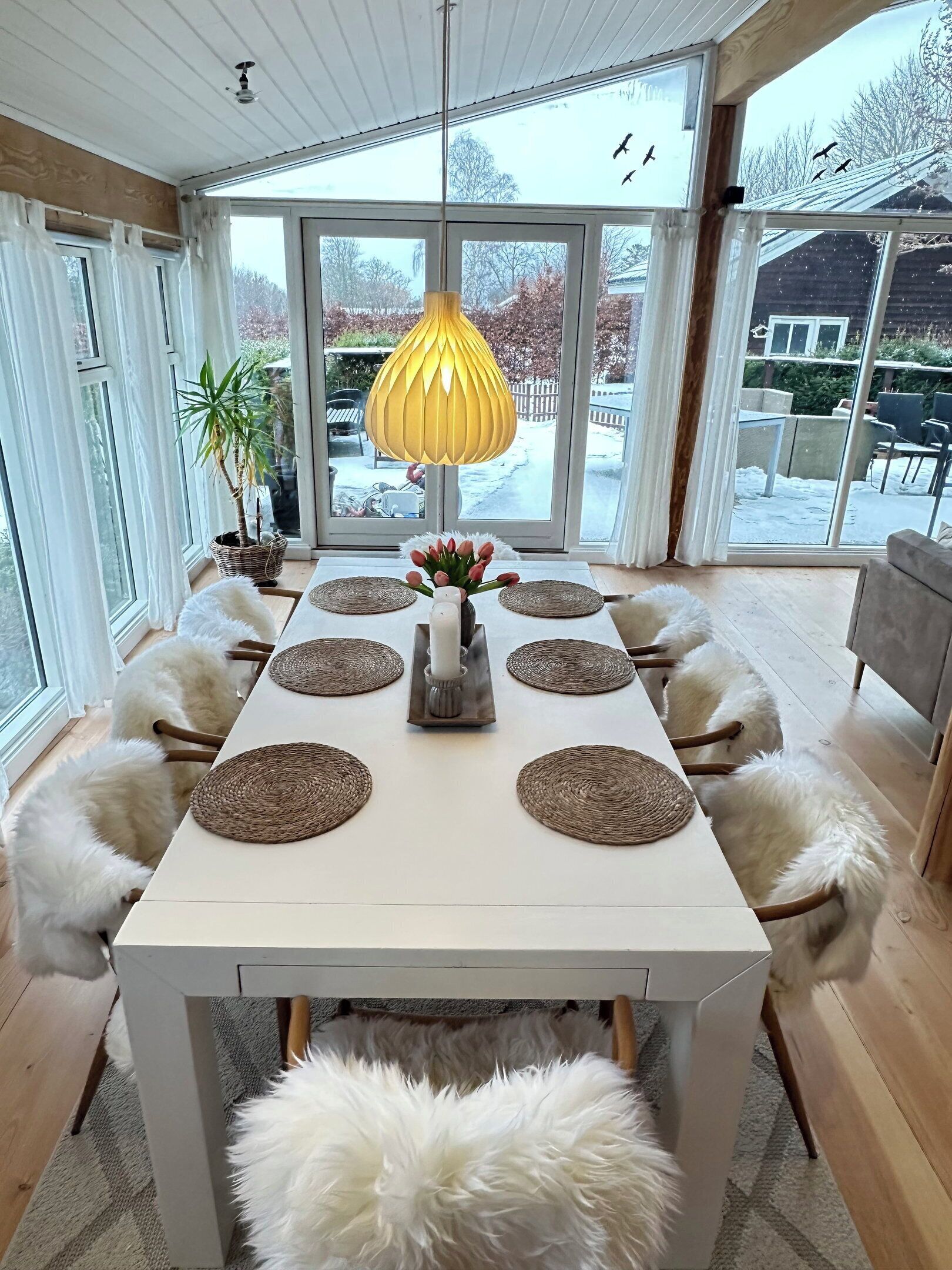 Un coin repas avec une table blanche, quatre chaises, un luminaire suspendu jaune et une vue sur l’extérieur.