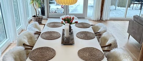 Un coin repas avec une table blanche, quatre chaises, un luminaire suspendu jaune et une vue sur l’extérieur.