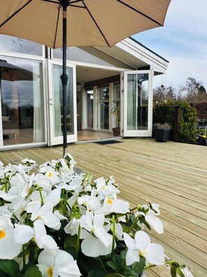 A modern house with large glass doors, a wooden deck, and white flowers in the foreground. - Spacious holiday home near beach and nature (Jægerspris)