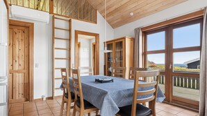 Un comedor con una mesa dispuesta para cuatro, sillas de madera, una escalera y vista al mar a través de un ventanal.