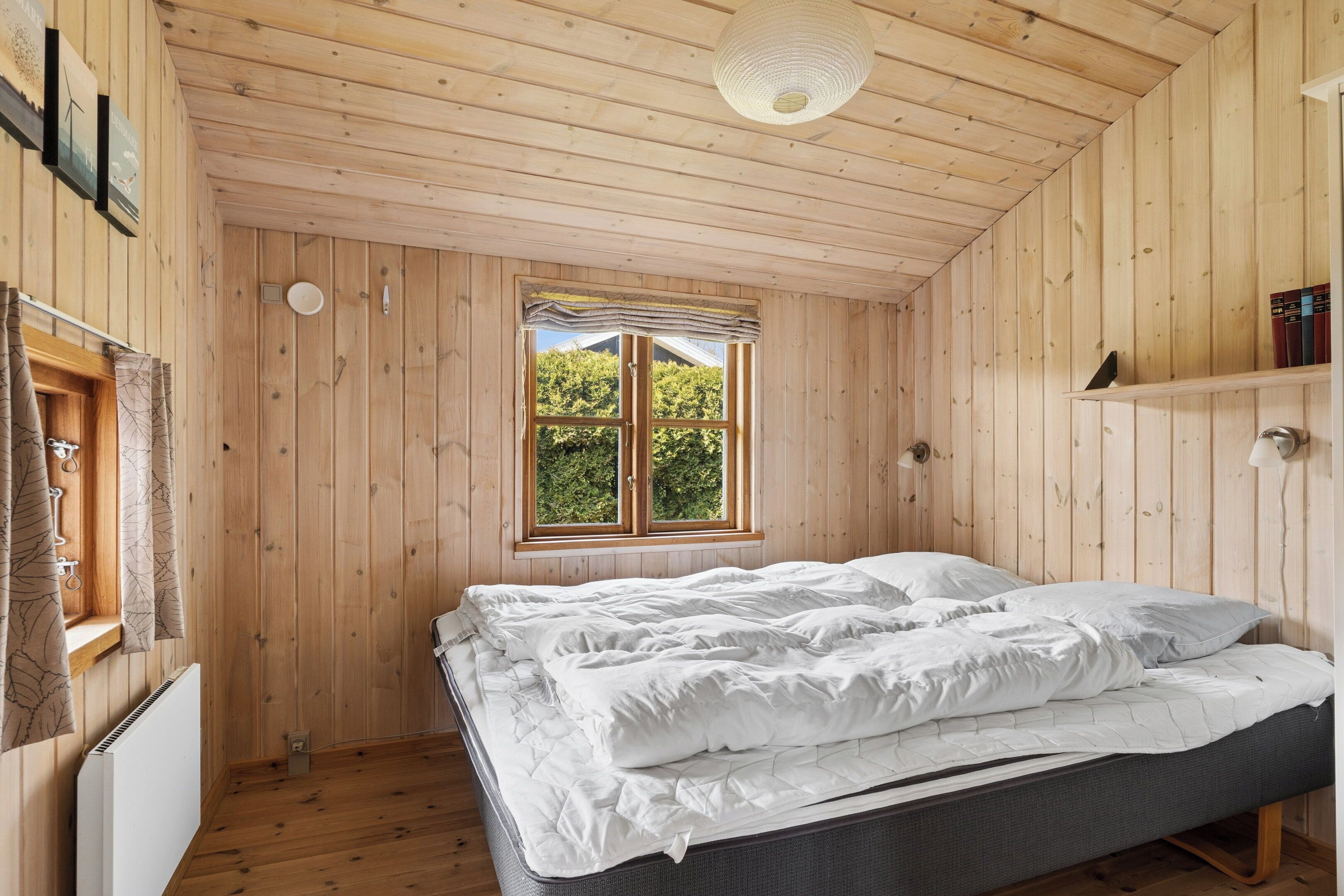 A wooden-paneled room with a bed, a window with curtains, and a shelf with books.